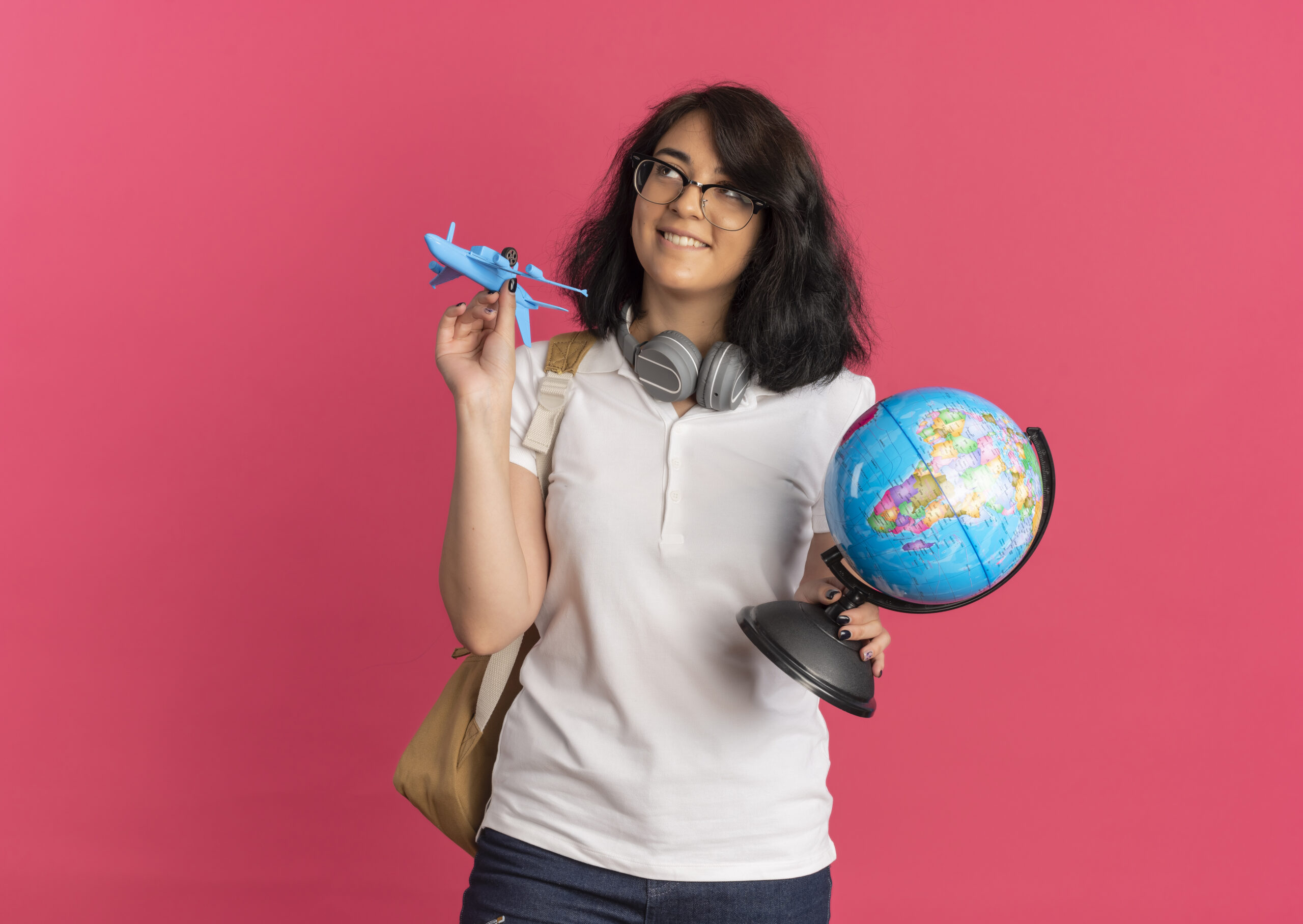 young pleased pretty caucasian schoolgirl with headphones on neck wearing glasses and back bag holds plane and globe looking up isolated on pink background with copy space