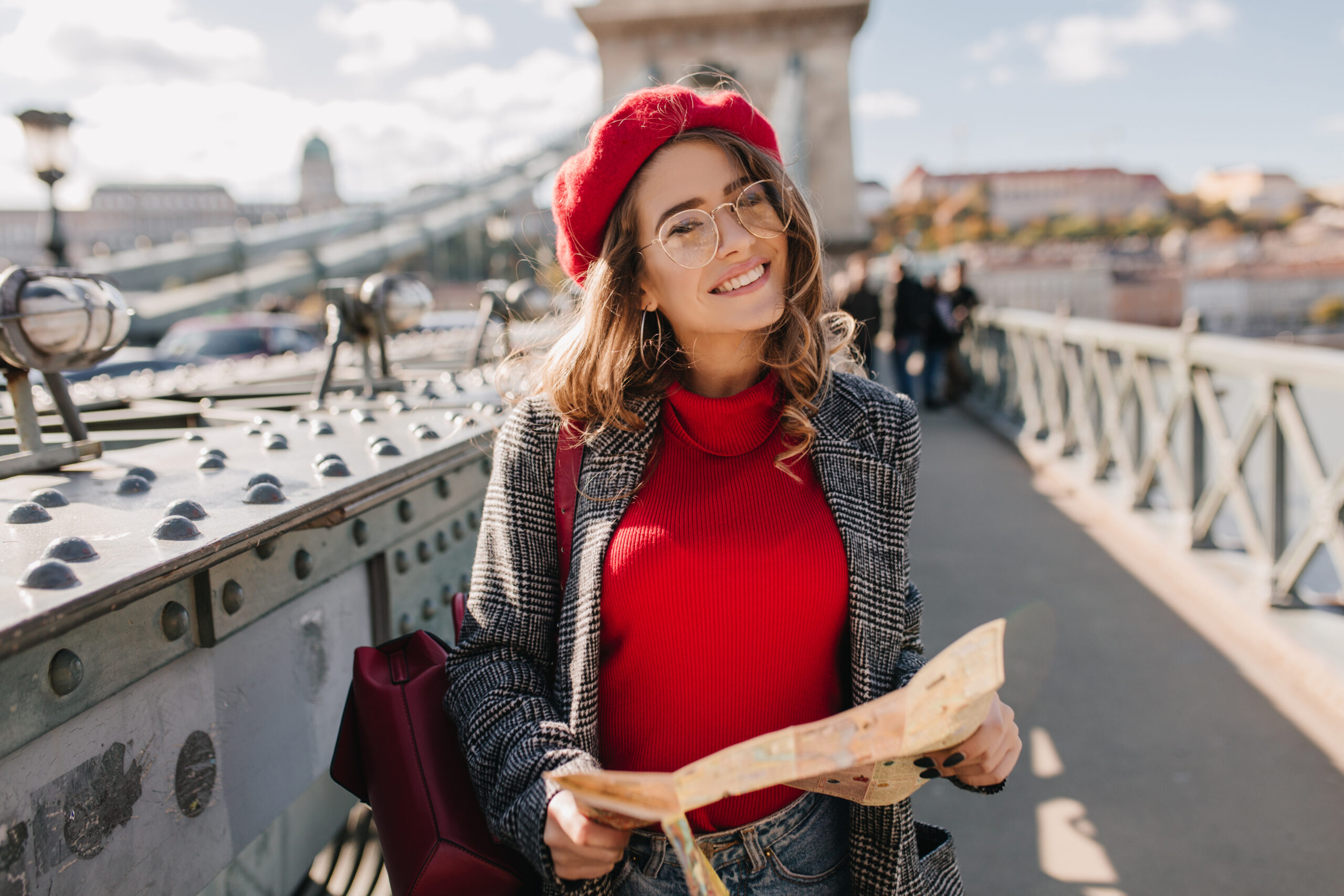 Pleased brunette woman with red backpack posing on bridge on urban background. Attractive white girl in glasses using map, exploring european city.. Pleased brunette woman with red backpack posing on bridge on urban background. Attractive white girl in glasses using map, exploring european city.